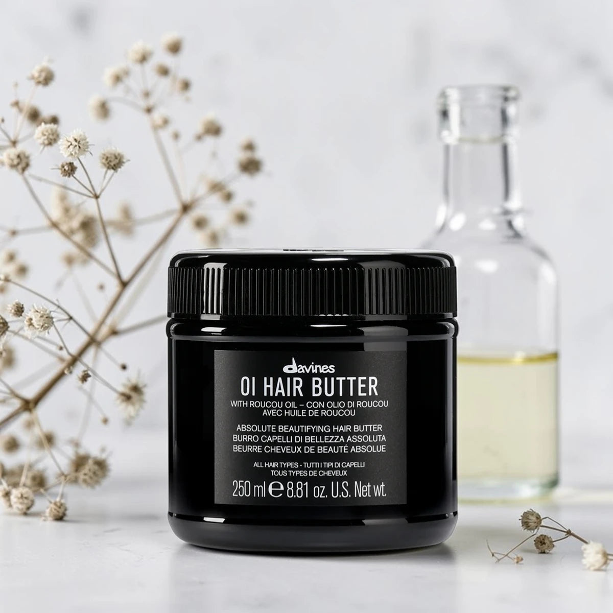 Pot noir de 250 ml du OI Hair Butter de Davines, baume de beauté absolue pour tous types de cheveux à l'huile de Roucou, posé aux côtés d'un flacon en verre et de fleurs séchées sur fond clair.