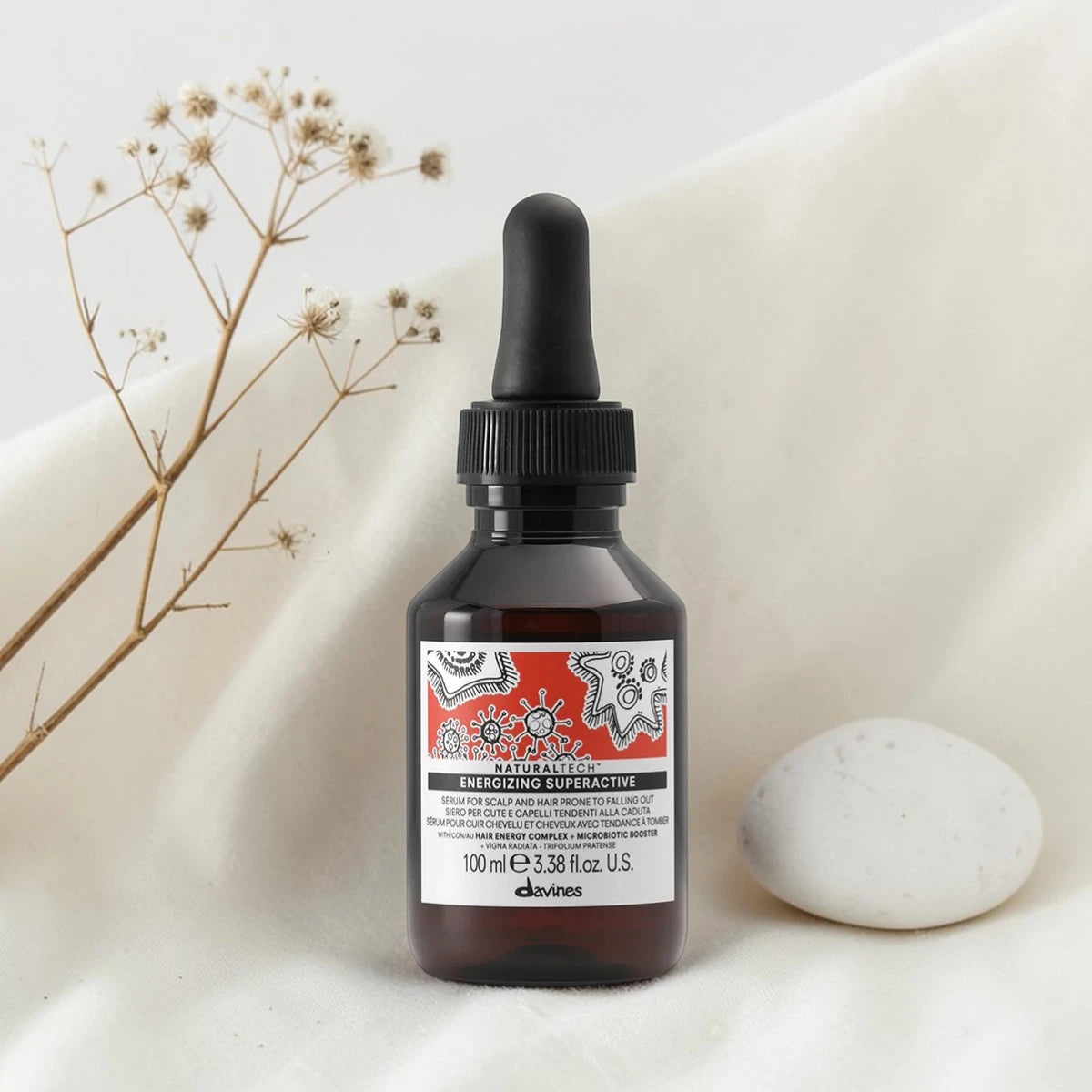 Flacon compte-gouttes de 100 ml du Naturaltech Energizing Superactive de Davines, sérum anti-chute pour cuir chevelu et cheveux fragiles au Hair Energy Complex et Microbiotic Booster, posé sur un tissu blanc aux côtés de fleurs séchées et d'un galet.