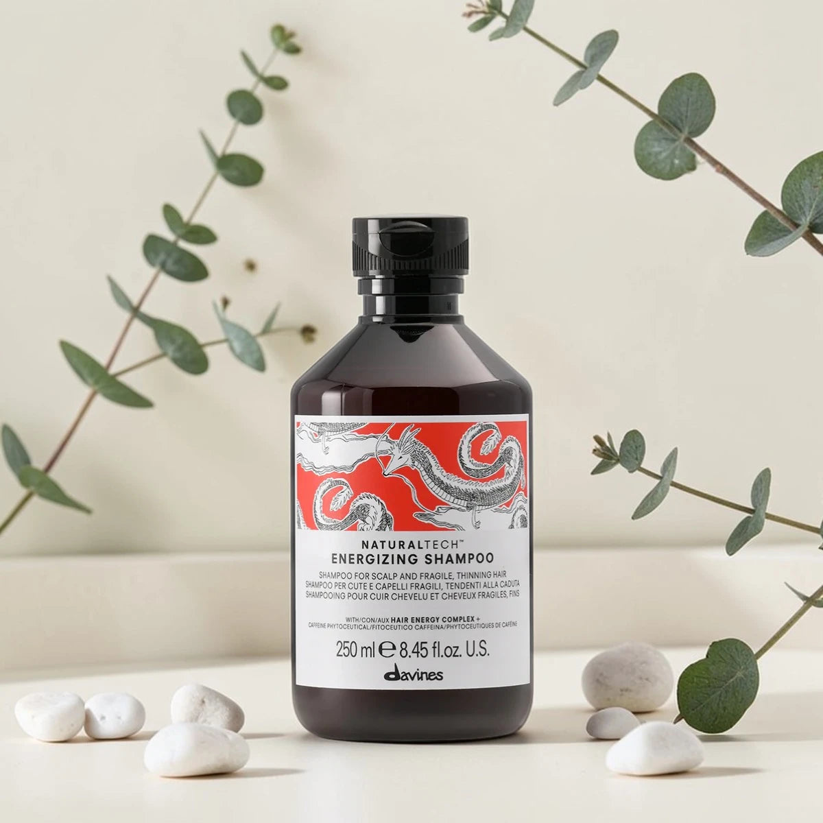 Flacon de 250 ml du Naturaltech Energizing Shampoo de Davines, shampooing anti-chute pour cuir chevelu et cheveux fragiles au Hair Energy Complex et aux phytoceutiques de caféine, entouré de feuilles d'eucalyptus et de galets blancs.