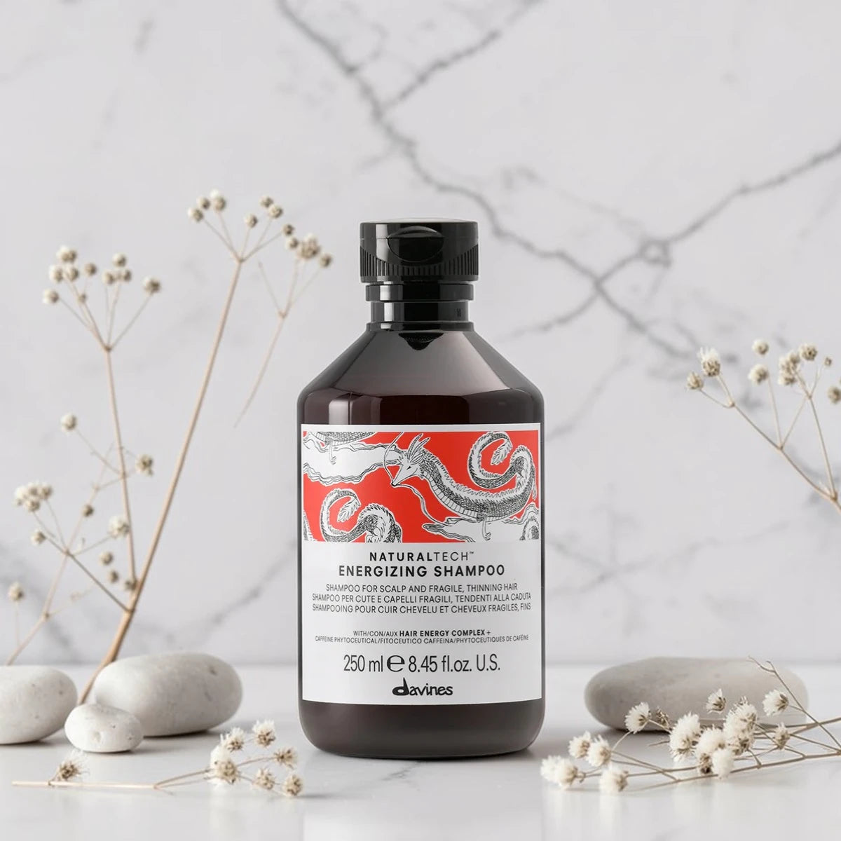 Flacon de 250 ml du Naturaltech Energizing Shampoo de Davines, shampooing anti-chute pour cuir chevelu et cheveux fragiles au Hair Energy Complex et aux phytoceutiques de caféine, posé sur un plan en marbre entouré de galets et de fleurs séchées.