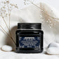 Pot noir de 250 ml du Heart of Glass Rich Conditioner de Davines, après-shampooing fortifiant et sublimateur pour cheveux blonds au Biacidic Bond Complex et à l'extrait de Jagua bleu, entouré de fleurs séchées et de galets blancs sur un tissu blanc.