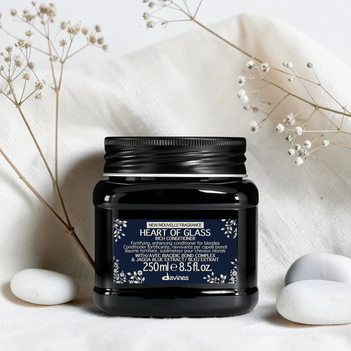 Pot noir de 250 ml du Heart of Glass Rich Conditioner de Davines, après-shampooing fortifiant et sublimateur pour cheveux blonds au Biacidic Bond Complex et à l'extrait de Jagua bleu, entouré de fleurs séchées et de galets blancs sur un tissu blanc.