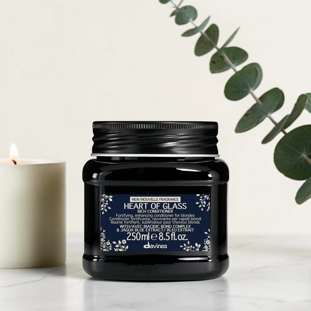 Pot noir de 250 ml du Heart of Glass Rich Conditioner de Davines, après-shampooing fortifiant et sublimateur pour cheveux blonds au Biacidic Bond Complex et à l'extrait de Jagua bleu, posé sur un plan en marbre aux côtés d'une bougie blanche et de feuilles d'eucalyptus.
