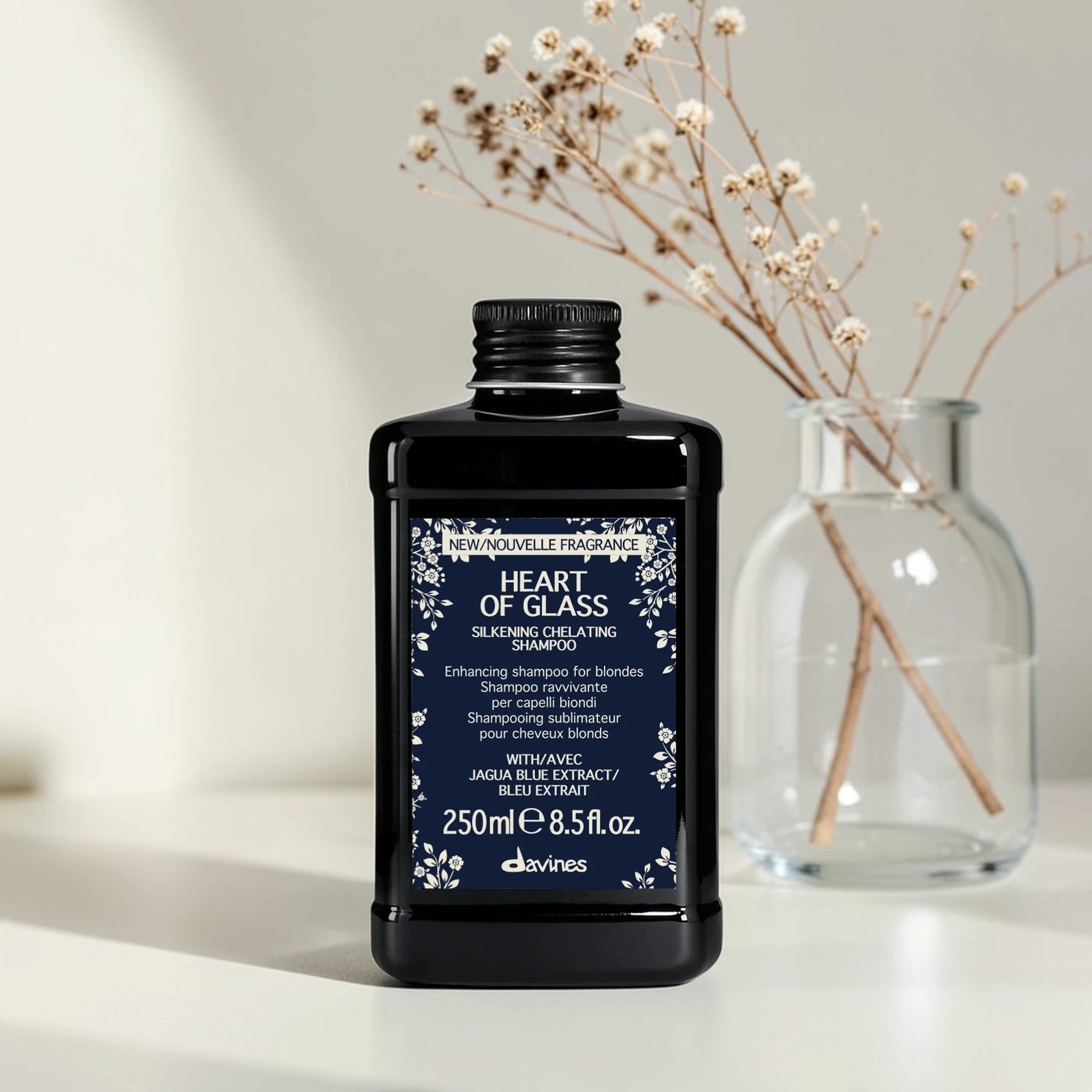 Flacon noir de 250 ml du Heart of Glass Silkening Chelating Shampoo de Davines, shampooing sublimateur pour cheveux blonds à l'extrait de Jagua bleu, posé sur une surface blanche aux côtés d'un vase en verre contenant des fleurs séchées.