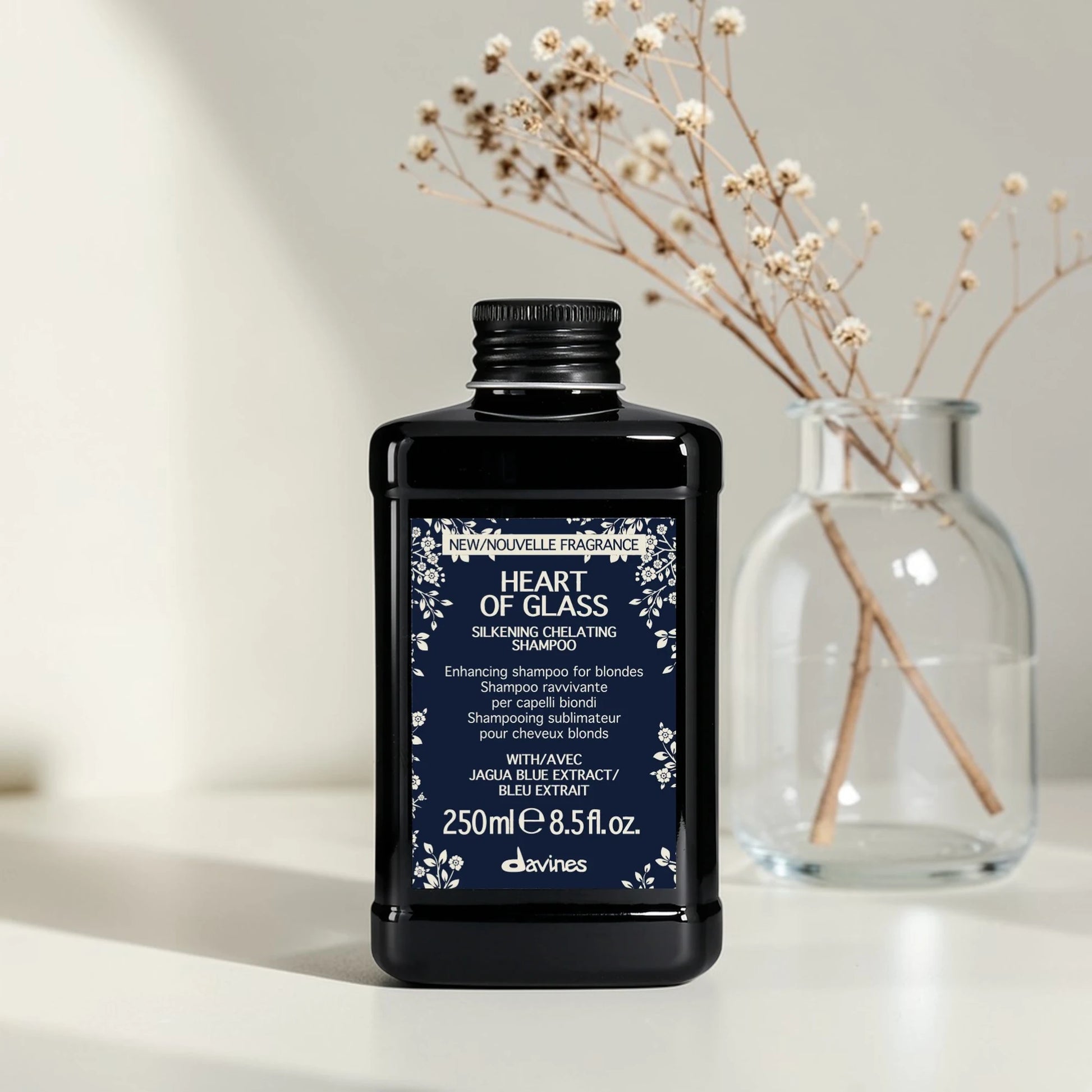 Flacon noir de 250 ml du Heart of Glass Silkening Chelating Shampoo de Davines, shampooing sublimateur pour cheveux blonds à l'extrait de Jagua bleu, posé sur une surface blanche aux côtés d'un vase en verre contenant des fleurs séchées.