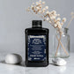 Flacon noir de 250 ml du Heart of Glass Silkening Chelating Shampoo de Davines, shampooing sublimateur pour cheveux blonds à l'extrait de Jagua bleu, posé sur un plan de travail en marbre aux côtés de galets et d'un vase contenant des fleurs séchées.