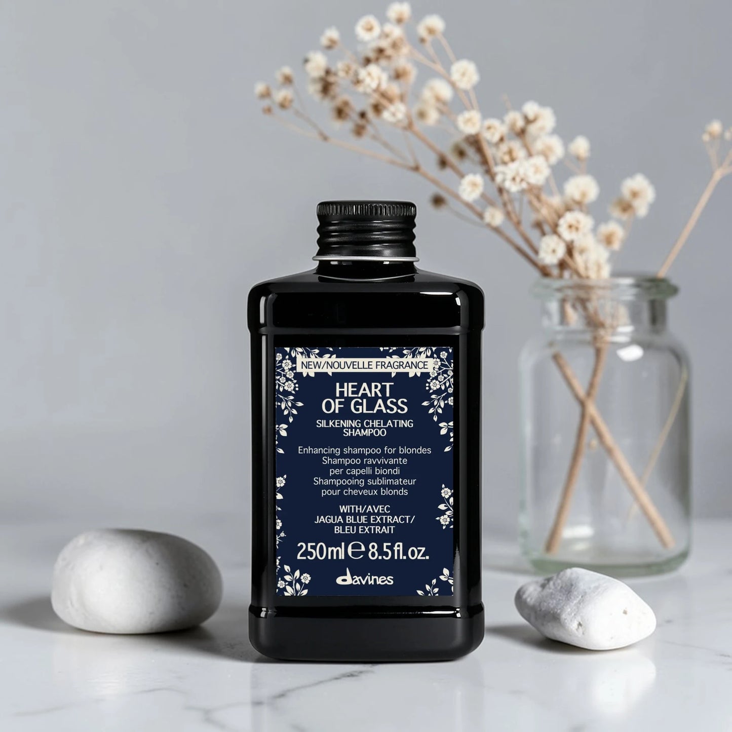 Flacon noir de 250 ml du Heart of Glass Silkening Chelating Shampoo de Davines, shampooing sublimateur pour cheveux blonds à l'extrait de Jagua bleu, posé sur un plan de travail en marbre aux côtés de galets et d'un vase contenant des fleurs séchées.