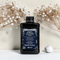 Flacon noir de 250 ml du Heart of Glass Silkening Chelating Shampoo de Davines, shampooing sublimateur pour cheveux blonds à l'extrait de Jagua bleu, entouré de fleurs séchées blanches et d'un galet.