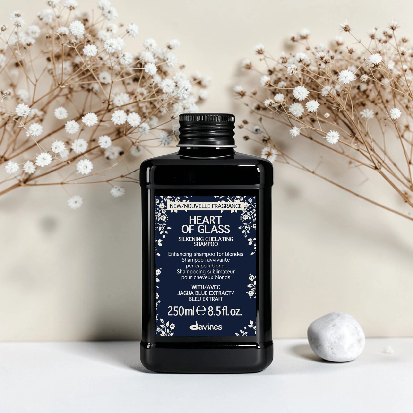 Flacon noir de 250 ml du Heart of Glass Silkening Chelating Shampoo de Davines, shampooing sublimateur pour cheveux blonds à l'extrait de Jagua bleu, entouré de fleurs séchées blanches et d'un galet.