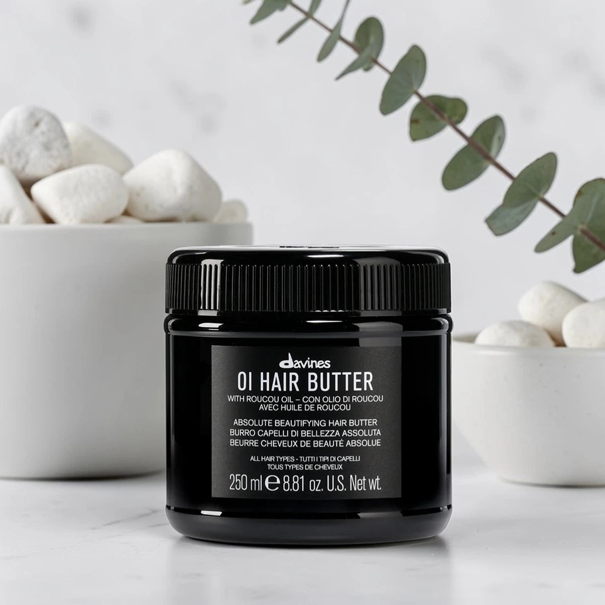 Pot noir de 250 ml du OI Hair Butter de Davines, baume de beauté absolue pour tous types de cheveux à l'huile de Roucou, posé sur un plan en marbre devant deux coupelles blanches remplies de galets et une branche d'eucalyptus.