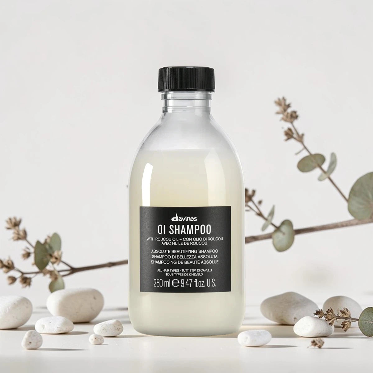 Flacon transparent de 280 ml du OI Shampoo de Davines, shampooing de beauté absolue pour tous types de cheveux à l'huile de Roucou, entouré de galets blancs et de branches d'eucalyptus.