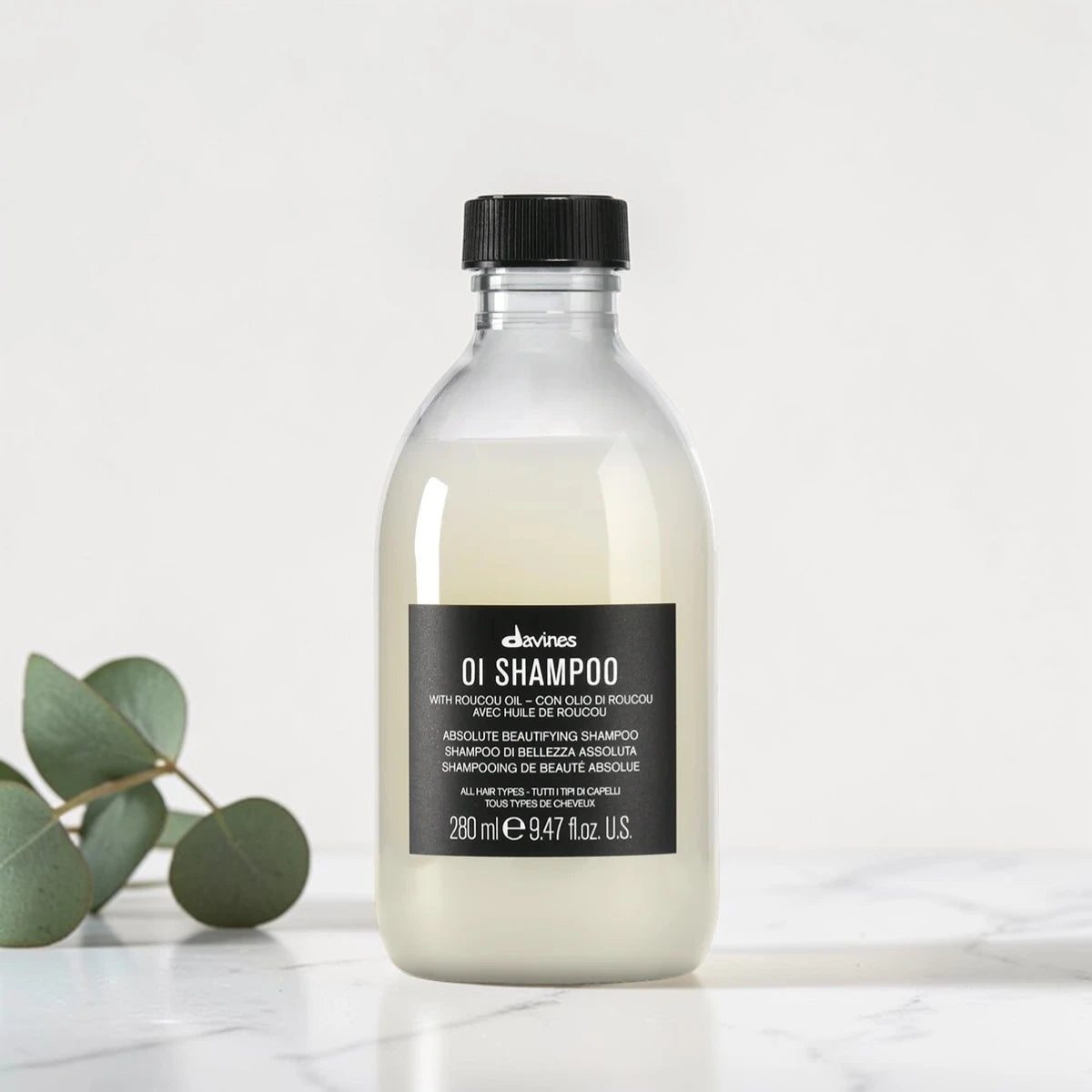 Flacon transparent de 280 ml du OI Shampoo de Davines, shampooing de beauté absolue pour tous types de cheveux à l'huile de Roucou, posé sur un plan en marbre aux côtés de feuilles d'eucalyptus.