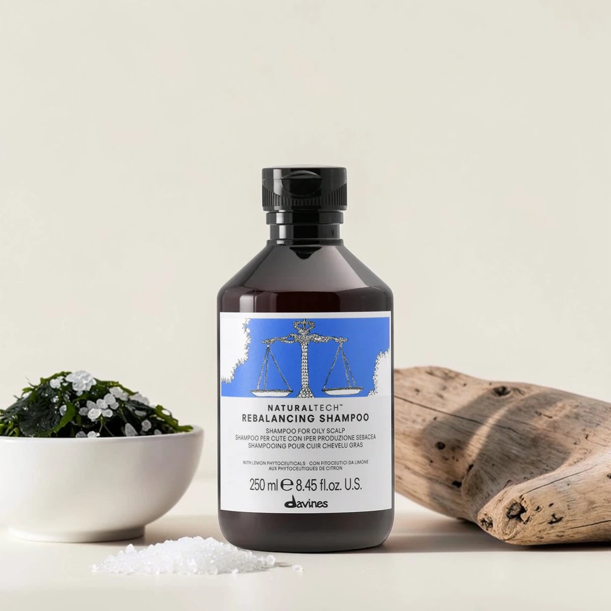 Flacon de 250 ml du Naturaltech Rebalancing Shampoo de Davines, shampooing pour cuir chevelu gras aux phytoceutiques de citron, posé aux côtés d'un morceau de bois flotté, d'un bol de plantes vertes et de cristaux de sel.