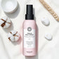 Bouteille rose de spray chauffant crème Maria Nila sur un fond blanc avec des fleurs en coton et un bol