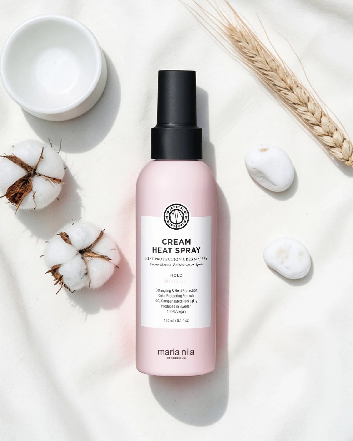 Bouteille rose de spray chauffant crème Maria Nila sur un fond blanc avec des fleurs en coton et un bol