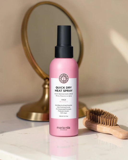 Flacon rose 'Quick Dry Heat Spray' avec capuchon noir sur une surface avec un miroir et une brosse en arrière-plan