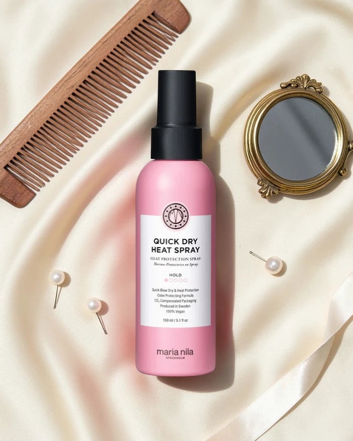 Flacon rose de Maria Nila Quick Dry Heat Spray sur un fond en tissu beige avec peigne et miroir