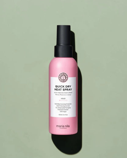 Flacon spray rose étiqueté "Quick Dry Heat Spray" par Maria Nila sur un fond vert clair