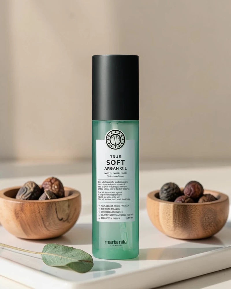 Bouteille d’huile d’argan True Soft de Maria Nila sur un fond neutre avec des bols en bois et des fruits d’argan