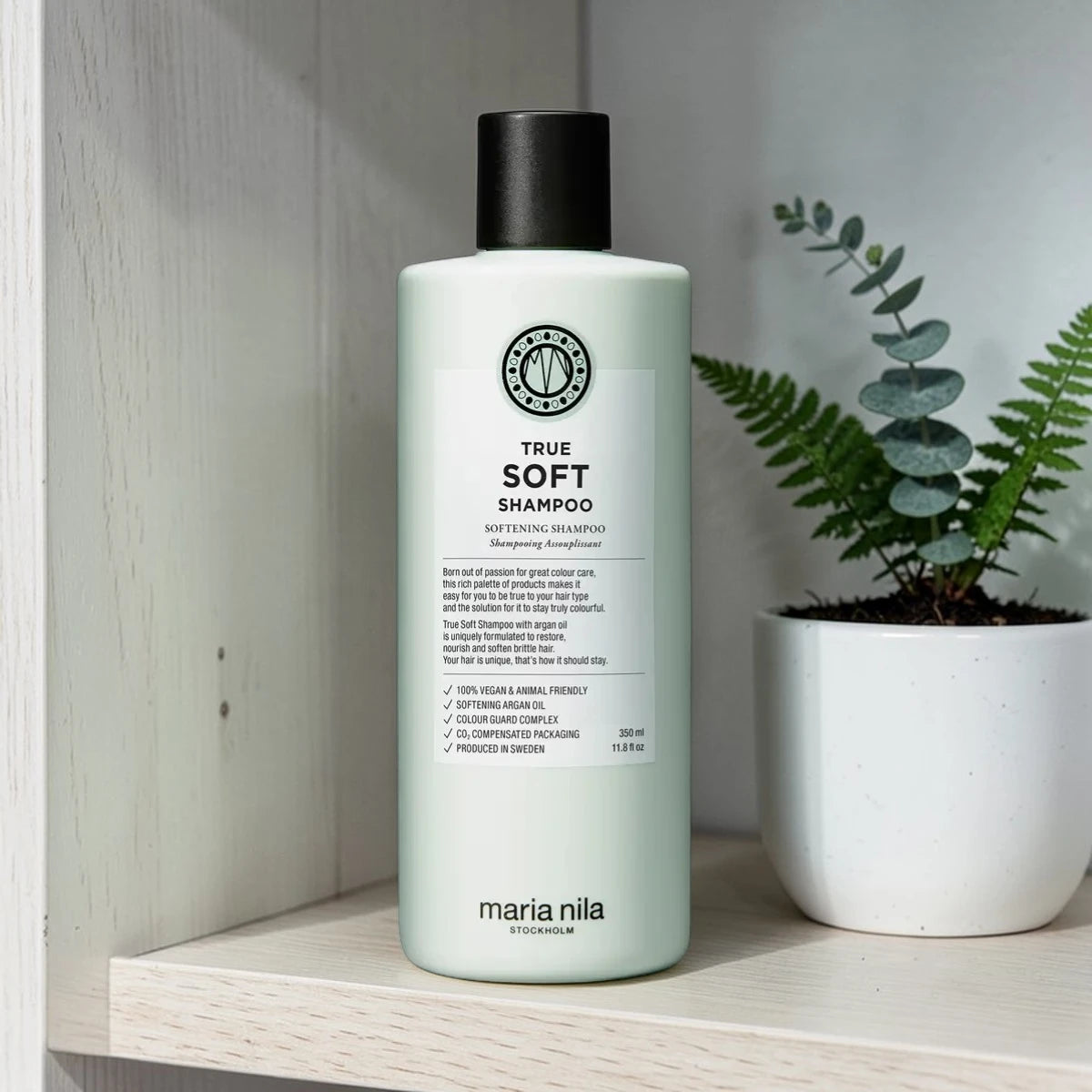 Bouteille de shampooing étiquetée 'True Soft Shampoo' par maria nila sur une étagère en bois avec une plante en arrière-plan