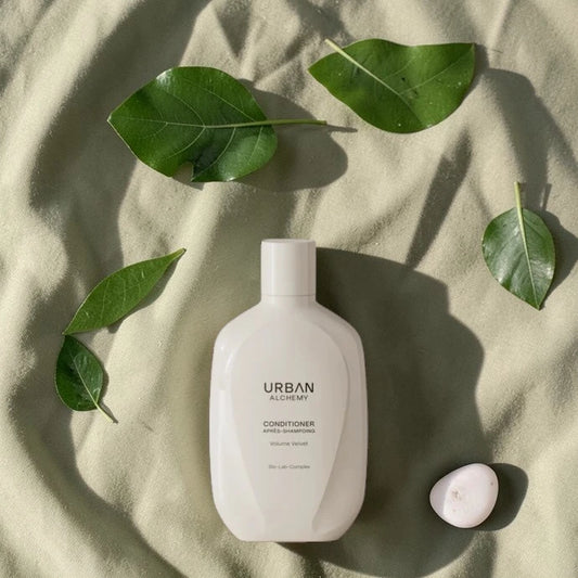 Bouteille blanche d’après-shampoing Urban Alchemy avec des feuilles vertes sur un fond en tissu beige