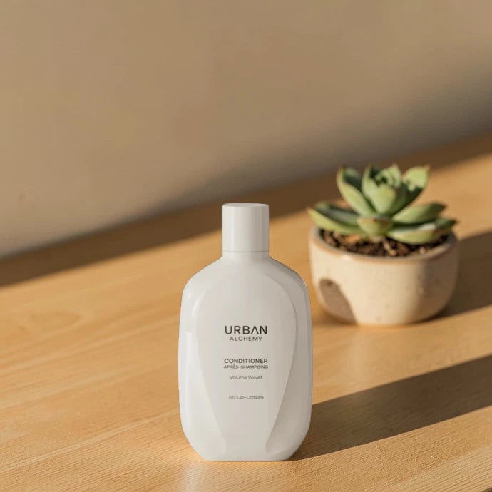 Bouteille blanche d’après-shampoing Urban Alchemy sur une surface en bois avec un pot de plante à l’arrière-plan